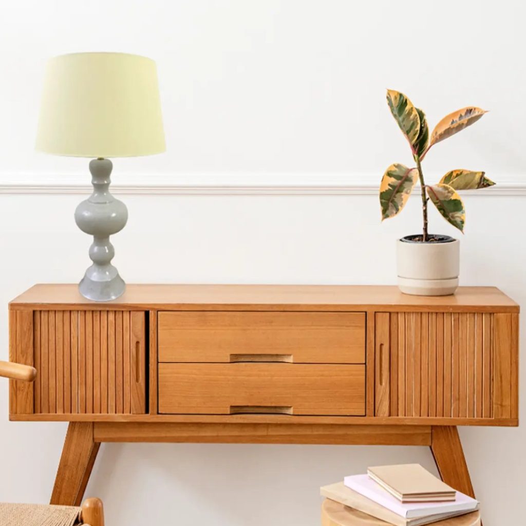 Lámpara de mesa con pantalla clara sobre mueble de madera junto a una planta en maceta.