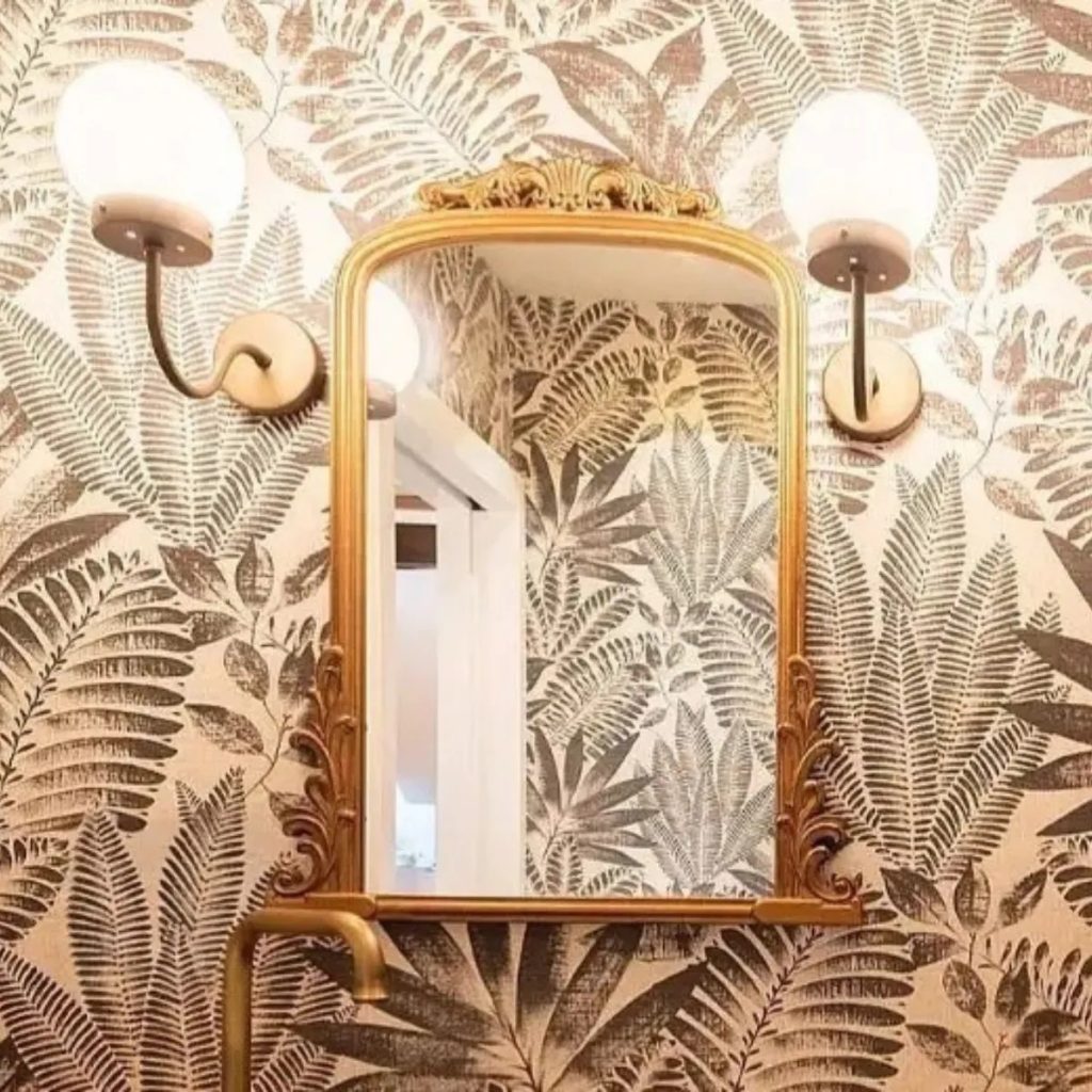Baño con espejo de marco dorado, apliques de pared con luz suave y papel pintado de hojas tropicales.