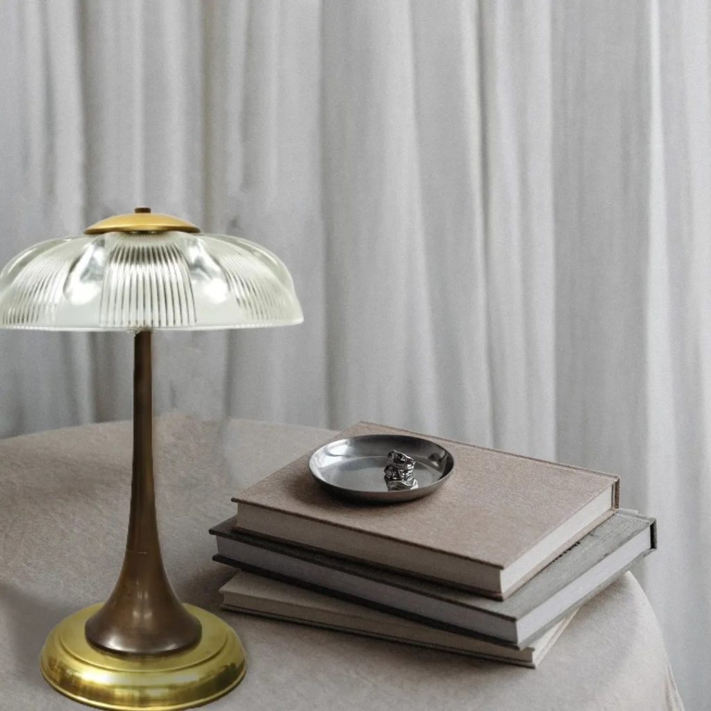Lámpara de mesa vintage con base metálica y pantalla de cristal estriado, colocada junto a libros en un ambiente minimalista.