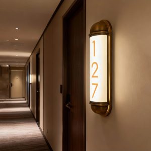 Aplique de pared vertical con iluminación integrada y número de habitación, instalado en el pasillo de un hotel.