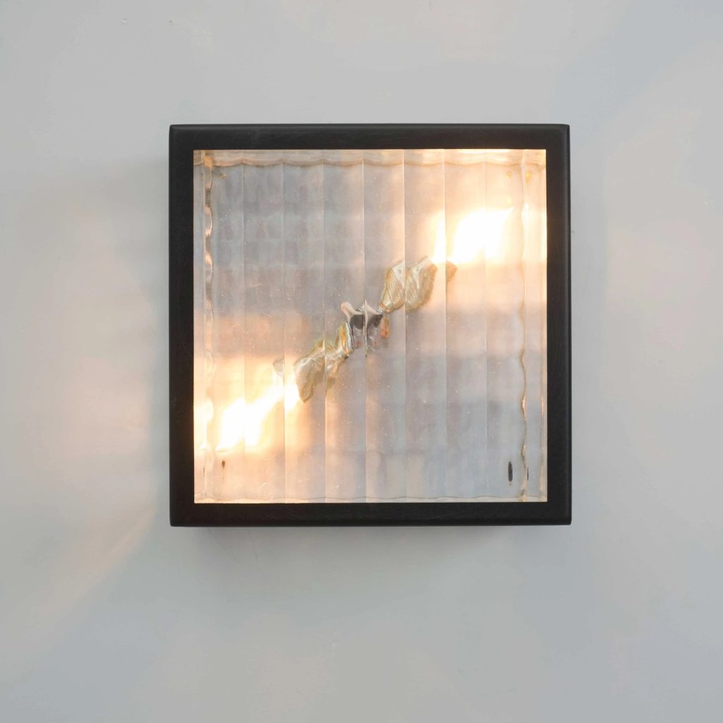 Aplique de pared cuadrado con marco negro y cristal texturizado iluminado en tono cálido.