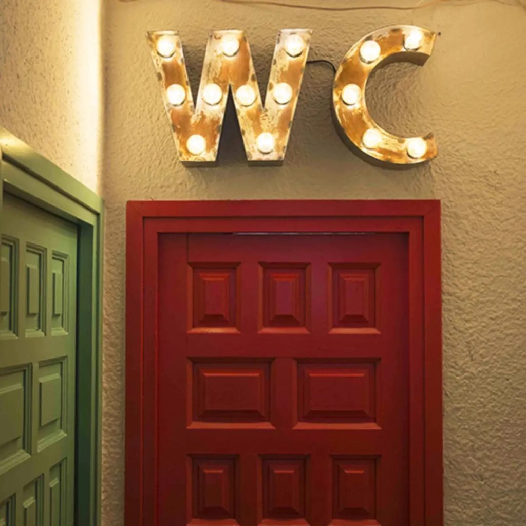 Cartel luminoso decorativo “WC” con bombillas vintage sobre una puerta roja de estilo retro industrial.