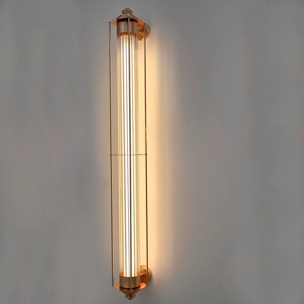 Aplique de pared vertical con tubo de cristal estriado y estructura en latón envejecido iluminado con luz cálida.
