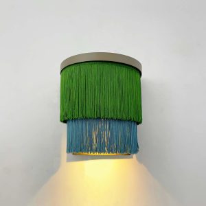 Lámpara de techo con pantalla de flecos verdes y azules, encendida sobre pared blanca.
