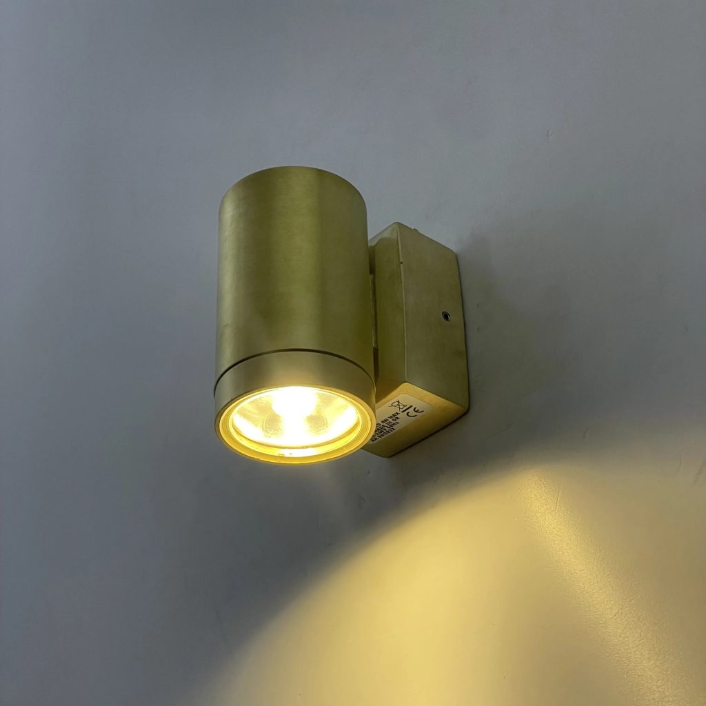 Aplique de pared cilíndrico en acabado dorado con foco LED orientado hacia abajo e iluminación cálida.