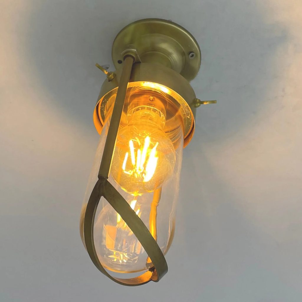 Plafón de techo vintage en latón envejecido con pantalla de cristal transparente y bombilla cálida vista.