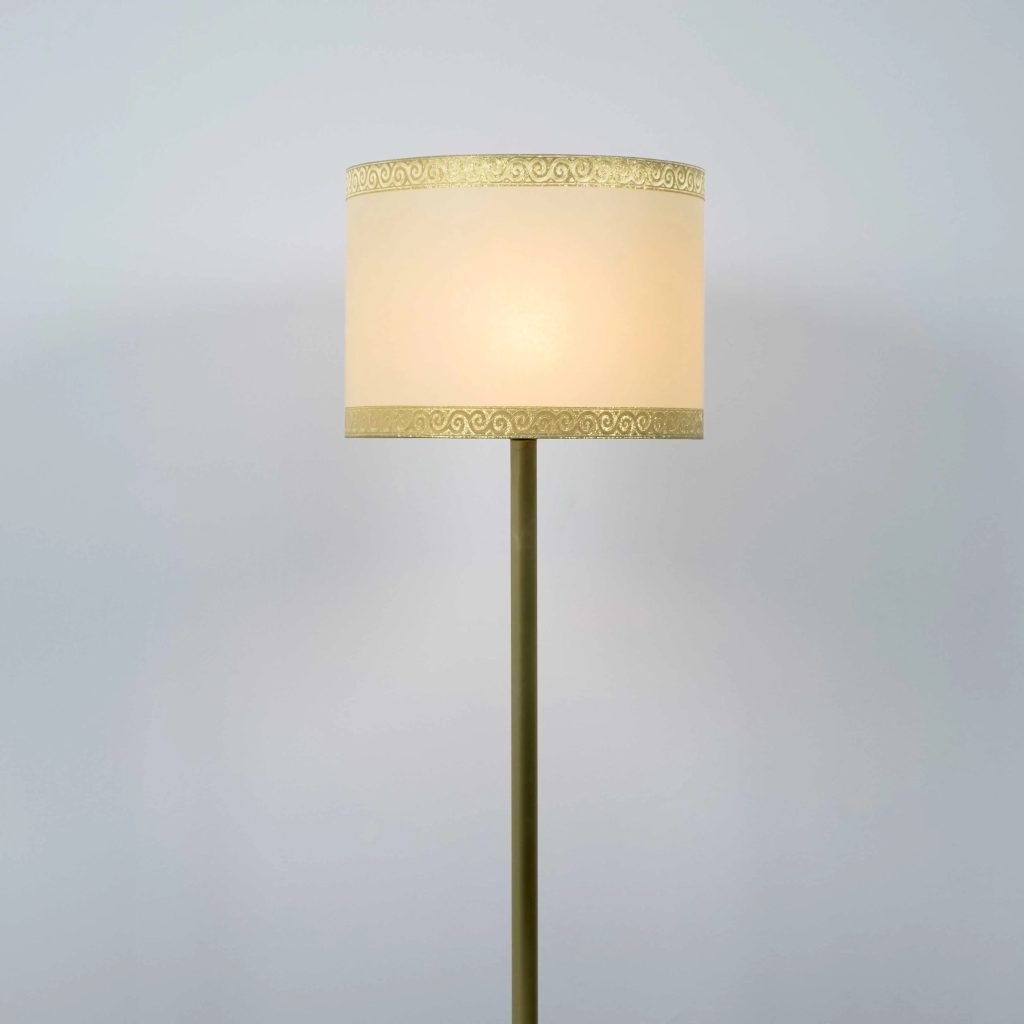 Lámpara de pie clásica con pantalla cilíndrica de tela crema y ribete decorativo dorado.