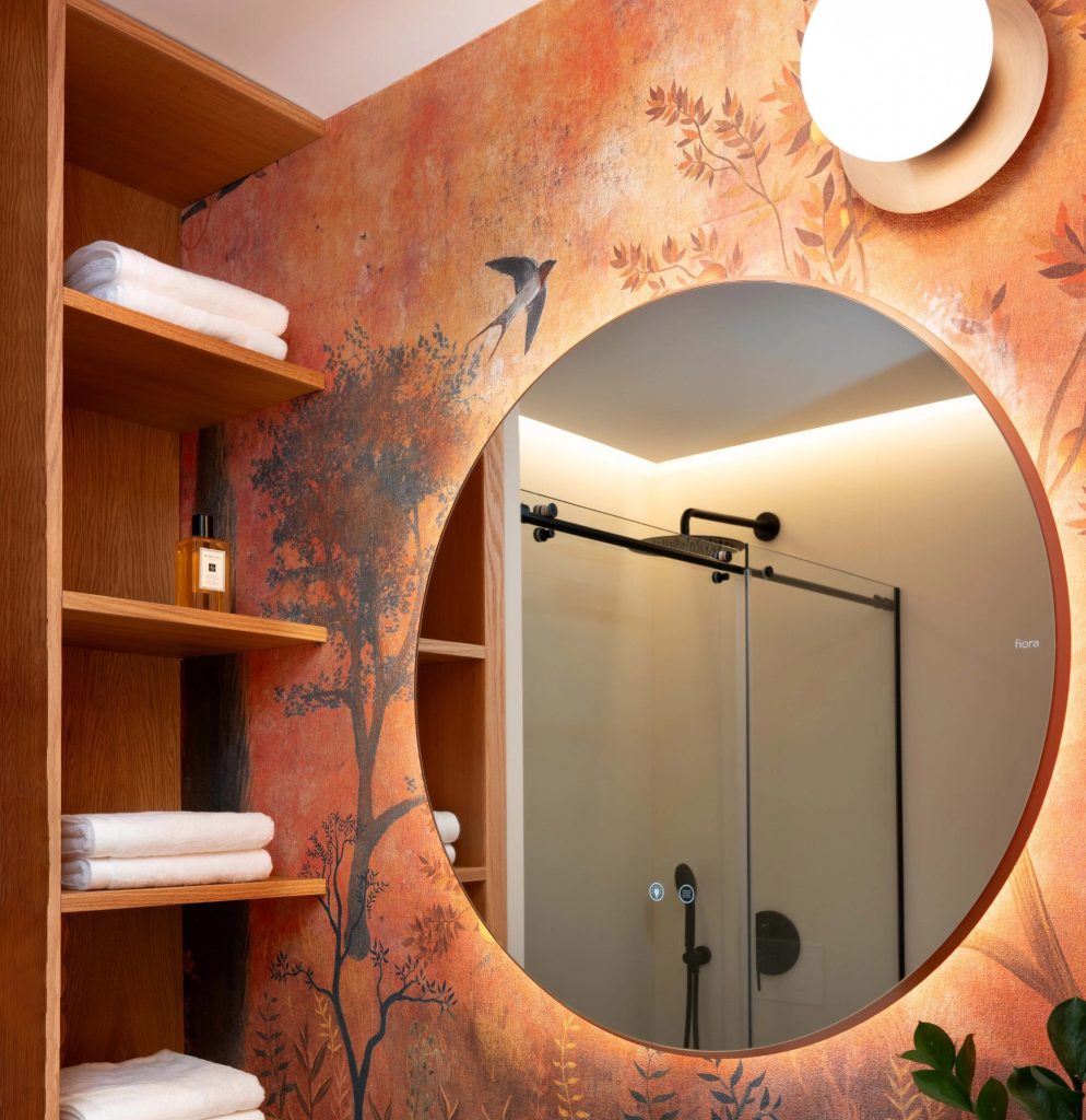 Aplique de pared con globo blanco instalado junto a espejo redondo en baño moderno con papel pintado decorativo.