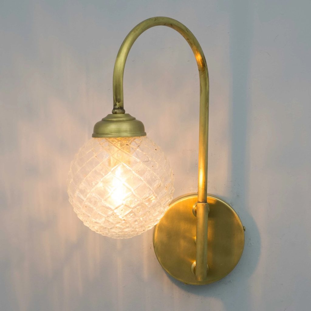 Aplique de pared dorado con brazo curvo y tulipa esférica de cristal tallado, encendido con luz cálida.