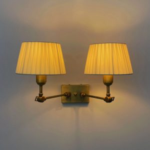 Aplique de pared doble en latón envejecido con pantallas plisadas de tela iluminadas con luz cálida.