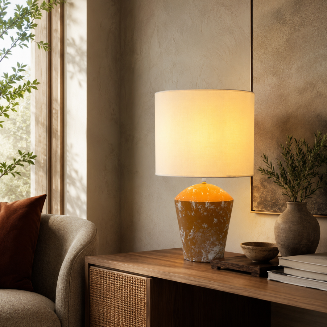 Lámpara de mesa con pantalla cilíndrica beige y base cerámica naranja jaspeada, encendida sobre un mueble de madera.