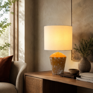 Lámpara de mesa con pantalla cilíndrica beige y base cerámica naranja jaspeada, encendida sobre un mueble de madera.