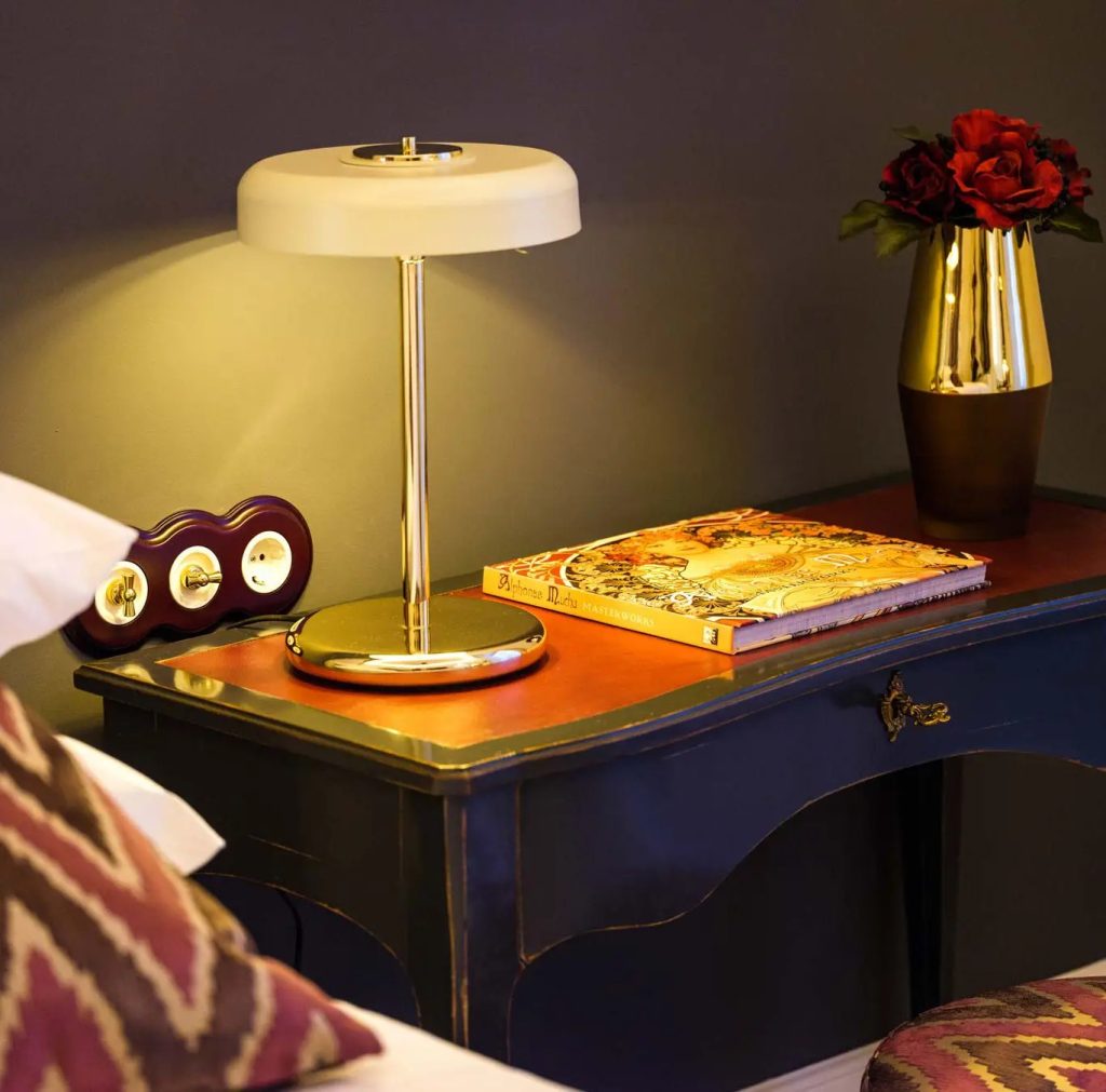 Lámpara de mesa vintage con pantalla metálica color crema, estructura de latón pulido y luz cálida sobre una mesita de noche.