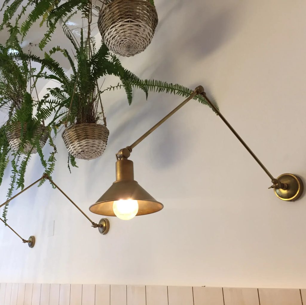 Aplique de pared articulado en acabado latón envejecido con pantalla cónica, instalado junto a plantas colgantes.