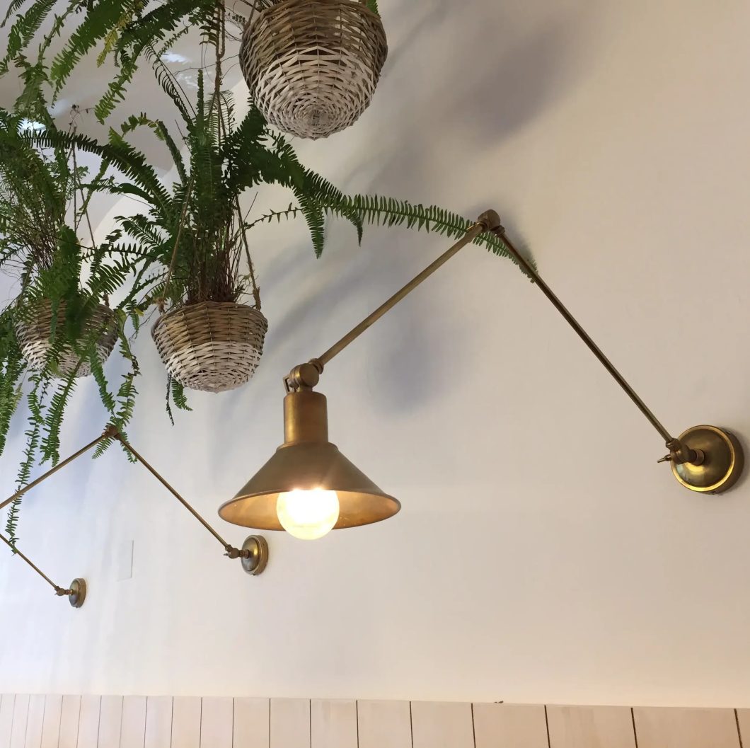 Aplique de pared articulado en acabado latón envejecido con pantalla cónica, instalado junto a plantas colgantes.