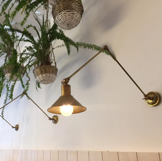 Aplique de pared articulado en acabado latón envejecido con pantalla cónica, instalado junto a plantas colgantes.