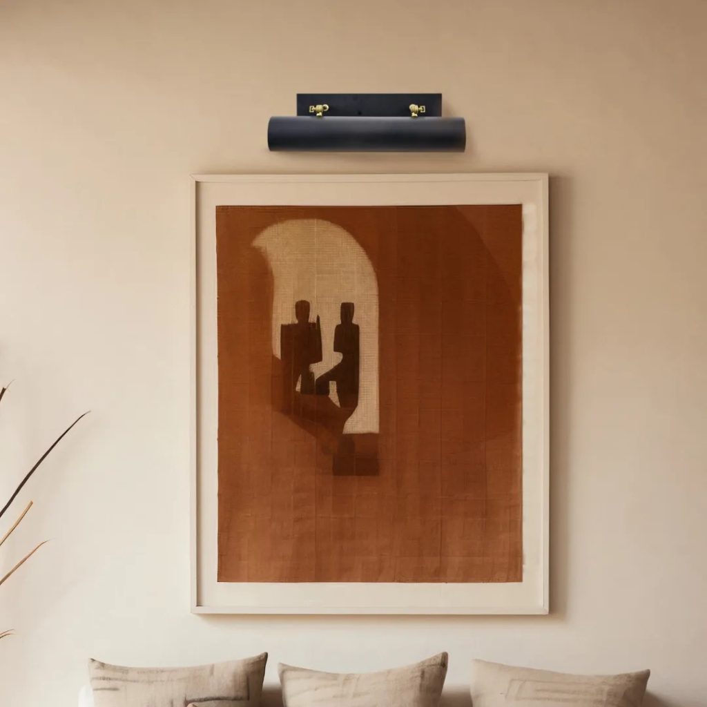 Aplique iluminacuadros negro con pantalla cilíndrica y detalles de latón instalado sobre una obra de arte.
