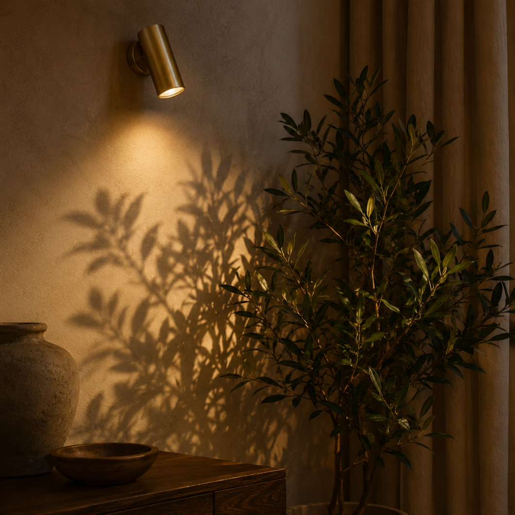 Foco de pared orientable de latón iluminando una planta decorativa y proyectando sombras sobre la pared.