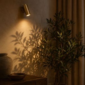 Foco de pared orientable de latón iluminando una planta decorativa y proyectando sombras sobre la pared.