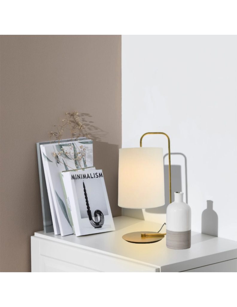 Lámpara de mesa de latón con pantalla de tela blanca encendida sobre una cómoda de estilo minimalista.