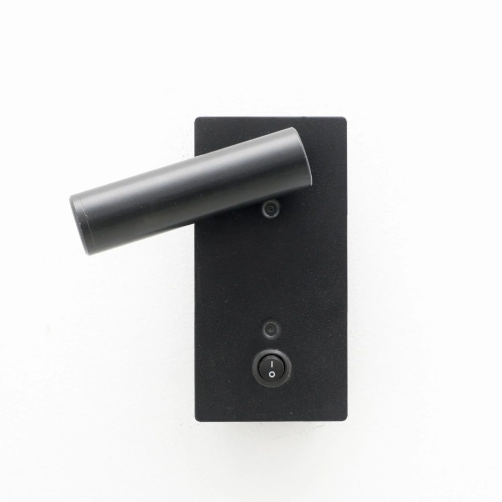 Aplique de pared negro con foco cilíndrico orientable y base rectangular, instalado sobre pared blanca.