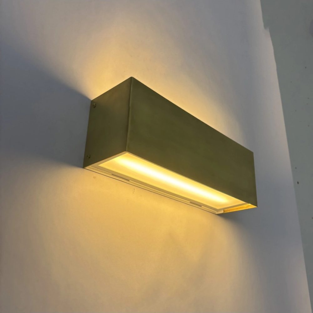 Aplique de pared rectangular en acabado dorado con luz LED cálida integrada, instalado sobre una pared blanca.