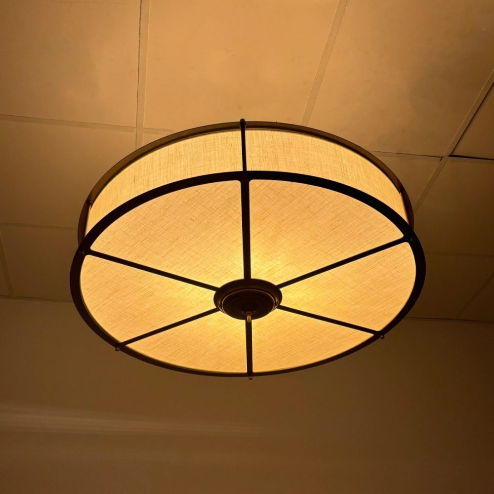 Plafón de techo circular con estructura metálica negra y pantalla de lino beige iluminada con luz cálida.